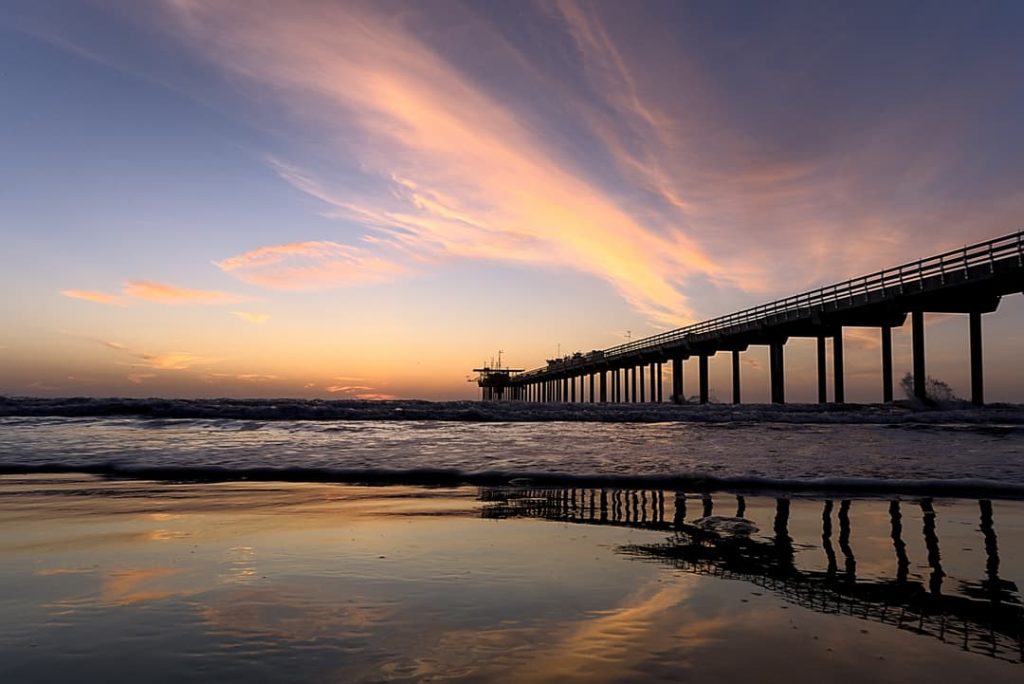 Een lange pier steekt de zee in terwijl de lucht boven San Diego oranje en roze kleurt bij zonsondergang, met golven die op het strand uitrollen