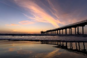Een lange pier steekt de zee in terwijl de lucht boven San Diego oranje en roze kleurt bij zonsondergang, met golven die op het strand uitrollen