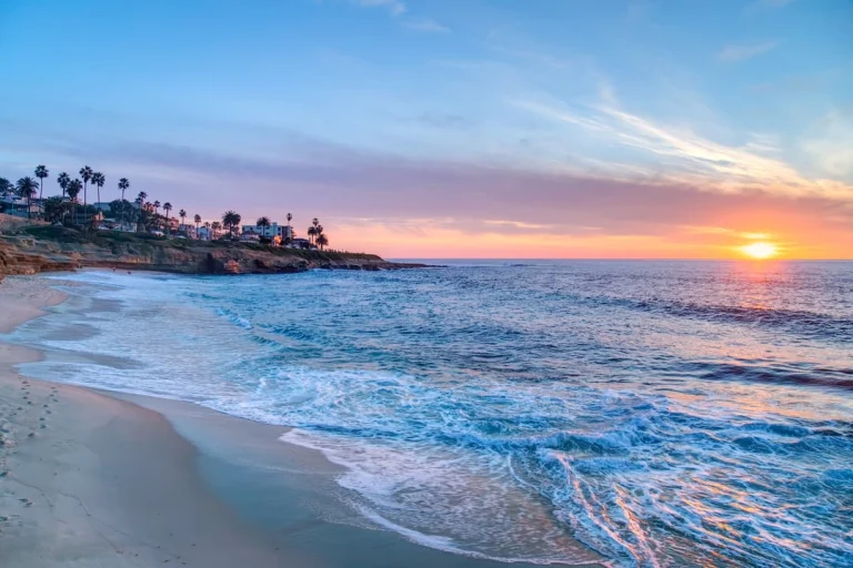 Golven spoelen over het zandstrand van San Diego terwijl de zon oranje kleurend achter de kustlijn met palmbomen ondergaat