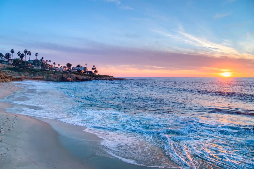 Golven spoelen over het zandstrand van San Diego terwijl de zon oranje kleurend achter de kustlijn met palmbomen ondergaat