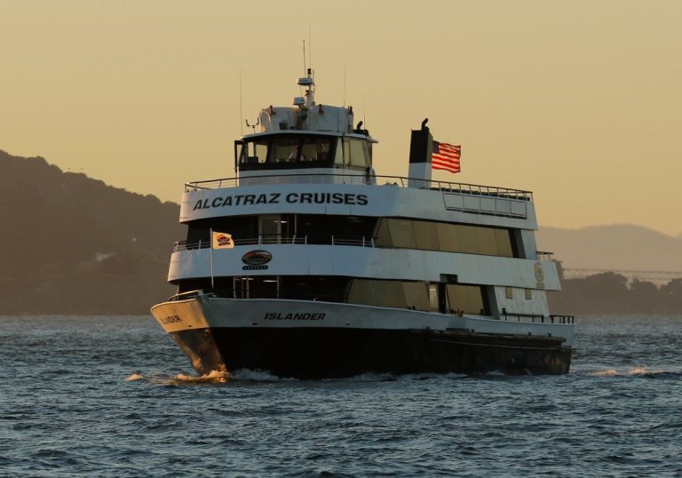 Een witte veerboot met opschrift Alcatraz Cruises en Amerikaanse vlag vaart over het water van San Francisco Bay tijdens zonsondergang