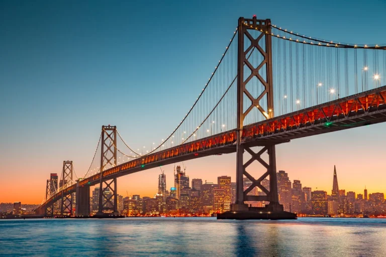 Verlichte hangbrug over het water met de skyline van San Francisco op de achtergrond bij zonsondergang