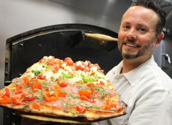 Lachende chef houdt een grote pizza met tomaat, kaas en kruiden vast bij een houtoven in de keuken