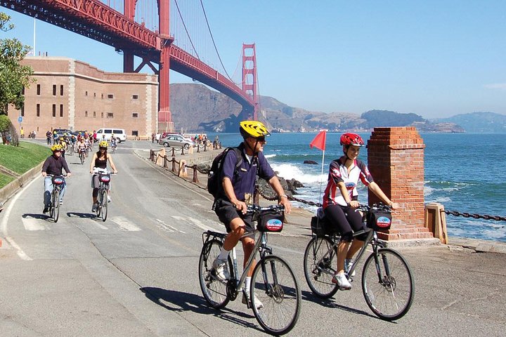 Fietsers met helmen rijden over een kustweg langs een bakstenen fort met de Golden Gate Bridge op de achtergrond