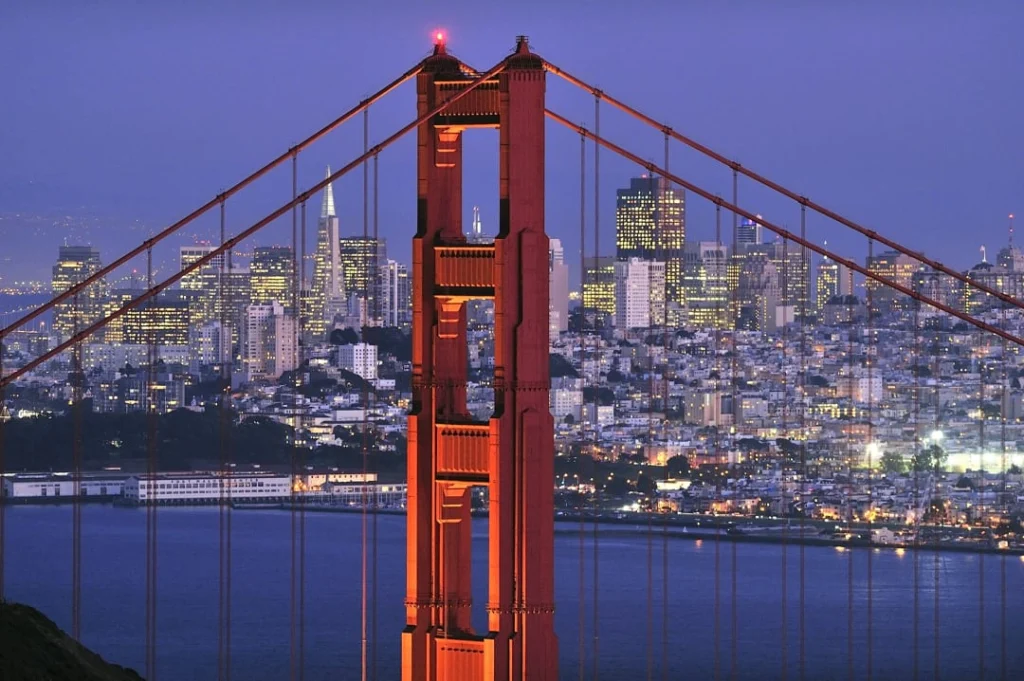 Rode brugtoren van de Golden Gate Bridge met de verlichte skyline van San Francisco op de achtergrond bij schemering