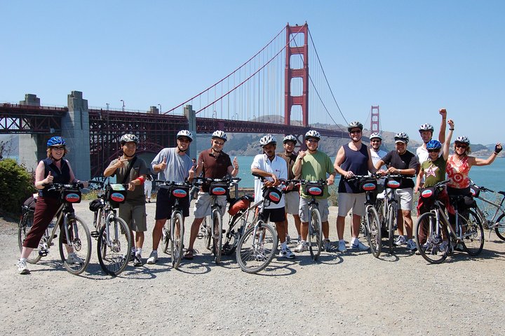 Grote groep fietsers met helmen staat met duimen omhoog voor de rode Golden Gate Bridge