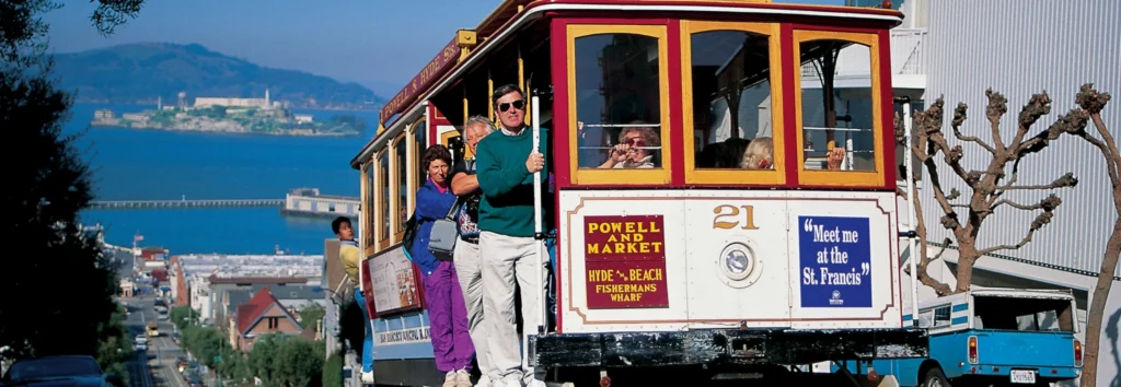 Toeristen op een historische kabeltram in San Francisco met de baai en het eiland Alcatraz op de achtergrond