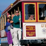 Toeristen op een historische kabeltram in San Francisco met de baai en het eiland Alcatraz op de achtergrond