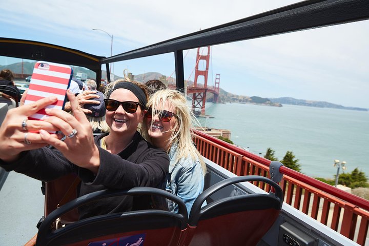 Twee lachende vrouwen maken een selfie op een open tourbus met de Golden Gate Bridge op de achtergrond