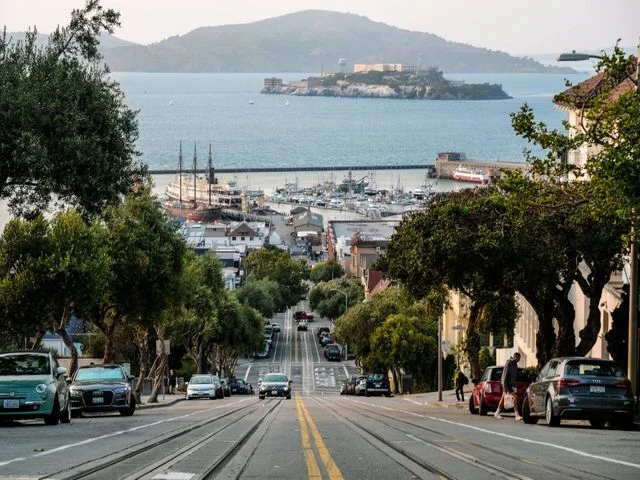 Steile straat met geparkeerde auto's loopt omlaag naar de baai, met Alcatraz eiland zichtbaar in de verte