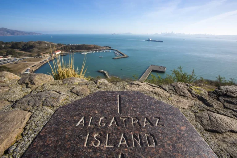 Stenen bord met opschrift Alcatraz Island op de voorgrond met uitzicht over de baai van San Francisco met een jachthaven en de skyline in de verte