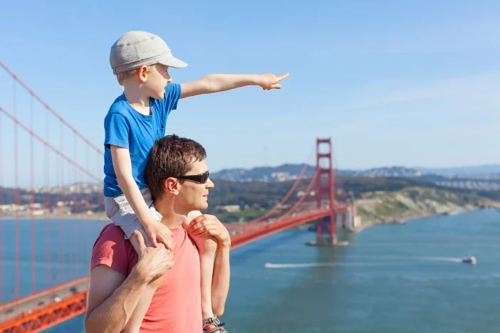 Vader draagt zijn zoontje op zijn schouders terwijl de jongen naar de rood oranje Golden Gate Bridge wijst