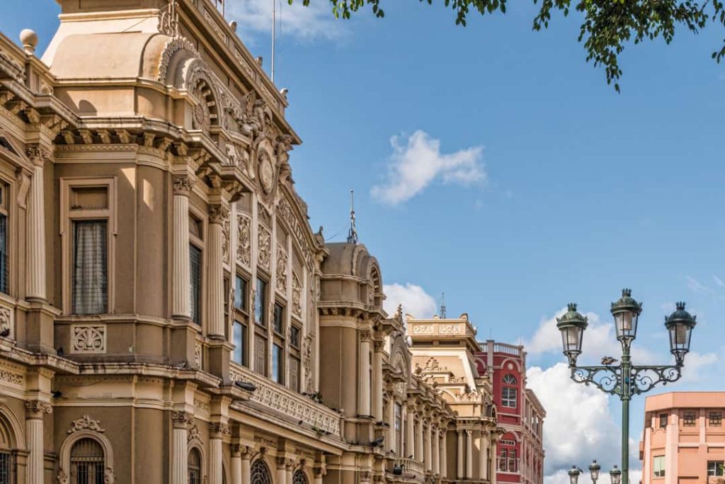 Sierlijke crèmekleurige gebouwgevel met versieringen en een zwarte lantaarnpaal op straat onder een blauwe lucht in San José