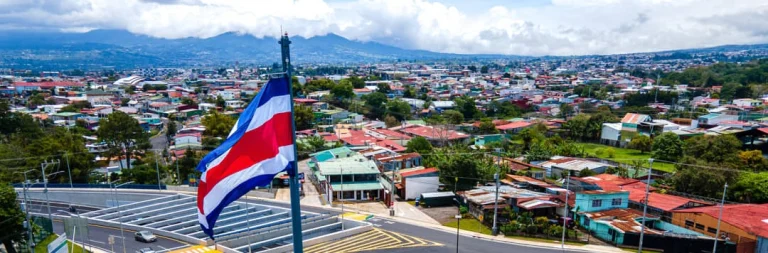 De rood wit blauwe vlag van Costa Rica wappert boven een uitgestrekte stad met kleurrijke daken en bergen op de achtergrond