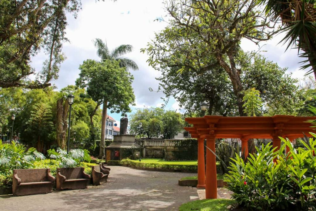 Groen stadspark met bankjes, een standbeeld en een oranje houten pergola op een zonnige dag in San José