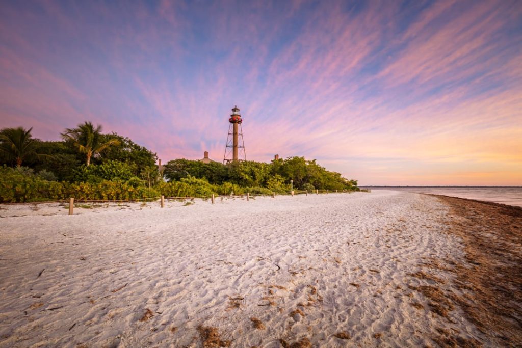 Silhouet van de vuurtoren van Sanibel Island tussen palmbomen op een leeg strand onder een roze en paarse avondlucht