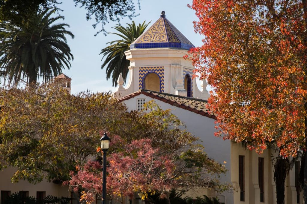 Sierlijke koepeltoren met blauwe en gele tegels van de Oude Missie van Santa Barbara steekt uit tussen palmbomen en rood herfstblad