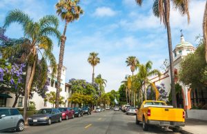 Rij geparkeerde auto's en een gele pick-up staan langs een straat vol hoge palmbomen met een wit gebouw met koepeltoren in Santa Barbara