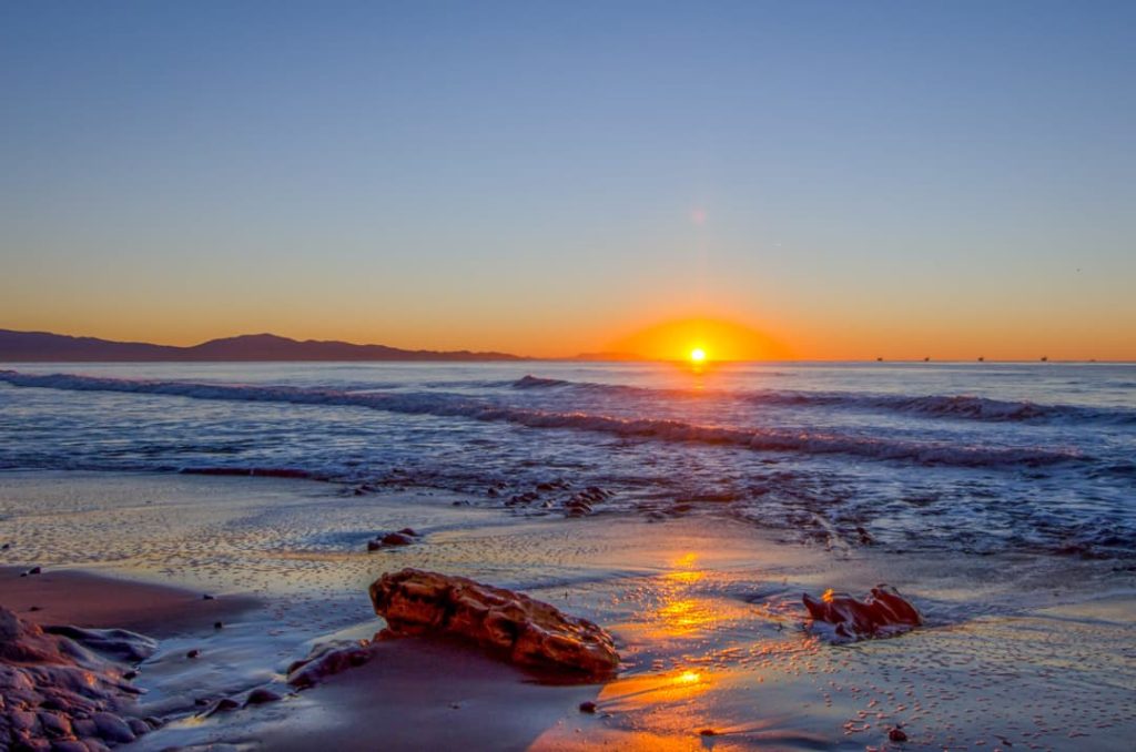 De ondergaande zon kleurt de lucht en het natte zand oranje terwijl golven over het strand bij Santa Barbara spoelen