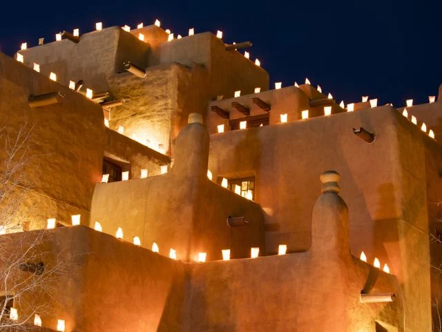 Terracottakleurig adobe gebouw in pueblostijl met tientallen kleine lichtjes op de daken en muren tegen een donkerblauwe avondlucht in Santa Fe
