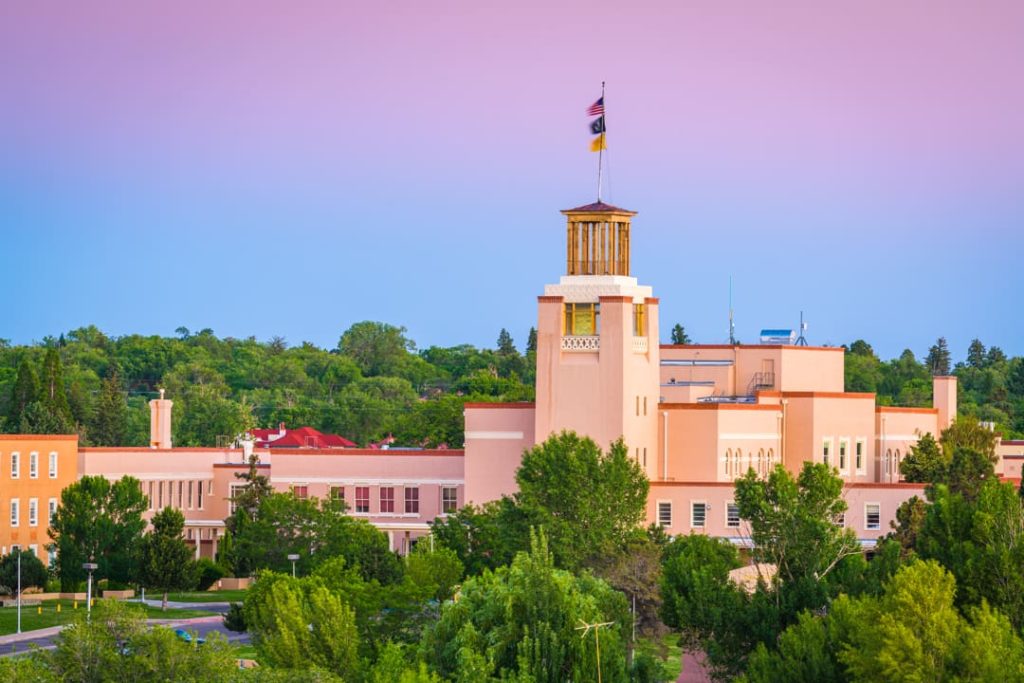 Roze pueblo-stijl gebouw met een houten uitkijktoren en een vlaggenmast, omgeven door bomen bij het vallen van de avond in Santa Fe