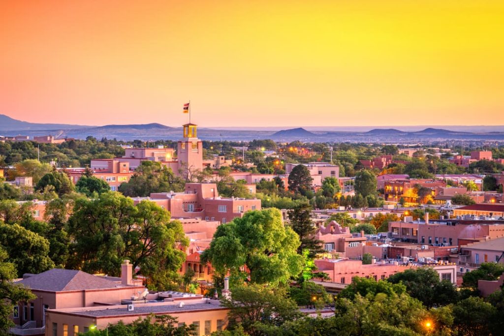 Luchtfoto van adobe gebouwen en een toren met vlaggen in Santa Fe, met bergen aan de horizon onder een felgele avondlucht