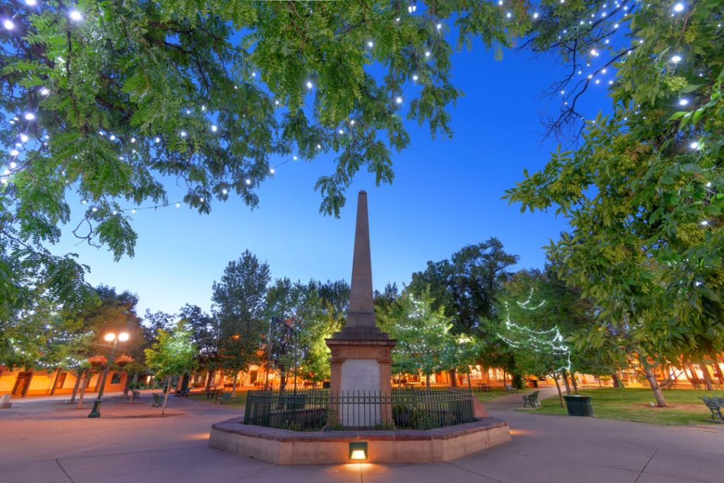 Stenen obelisk op een pleintje omgeven door bomen vol lichtjes en straatlantaarns in het centrum van Santa Fe bij het vallen van de avond