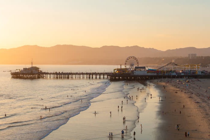 Luchtfoto van de houten pier van Santa Monica met een verlicht pretpark en reuzenrad, een breed zandstrand en de kustlijn bij zonsondergang