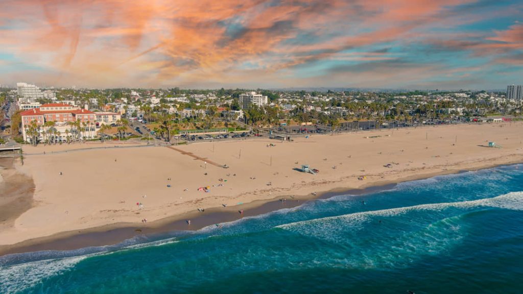 Luchtfoto van het brede zandstrand van Santa Monica met golvend water, palmbomen langs de boulevard en gebouwen in een roze avondlicht