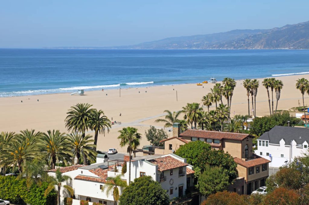 Uitzicht over palmbomen en huizen met terracotta daken naar het brede zandstrand en de blauwe oceaan van Santa Monica met bergen op de achtergrond