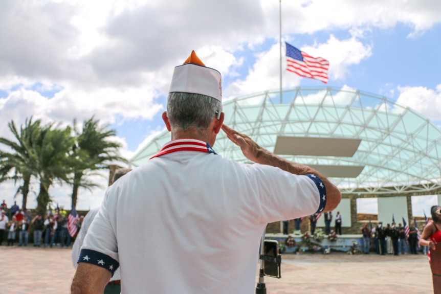 Man in wit uniform met puntmuts salueert naar een wapperende Amerikaanse vlag tijdens een herdenkingsceremonie op een begraafplaats