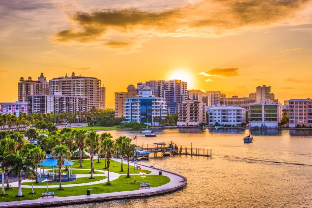Wolkenkrabbers van Sarasota kleuren goud bij zonsondergang met een park vol palmbomen en bootjes in de baai op de voorgrond