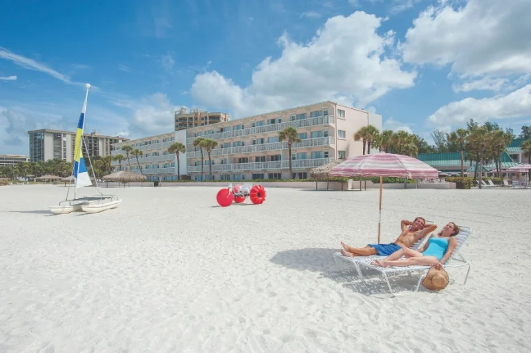 Een stel ligt te ontspannen op strandstoelen onder een parasol op een wit zandstrand in Sarasota met een hotel op de achtergrond
