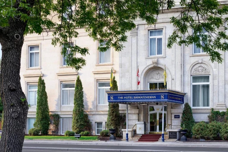 Voorgevel van het historische The Hotel Saskatchewan met een blauwe luifel boven de ingang en een boom ervoor op een heldere dag