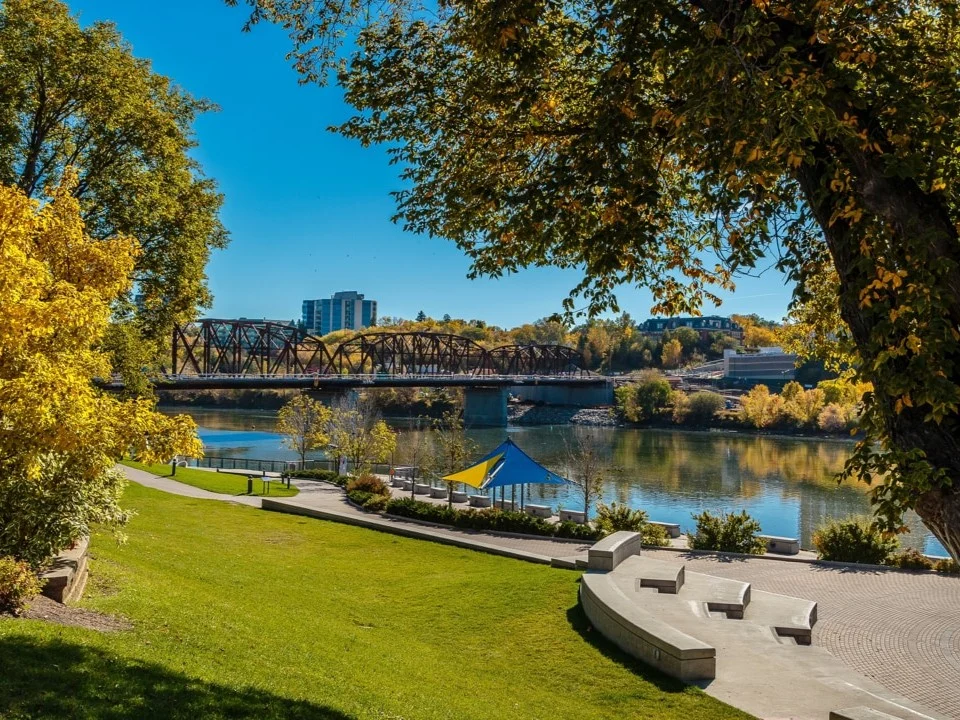 Wandelpad langs de rivier in Saskatoon met een gele driehoekige zonneschaduw, bankjes en herfstbladeren aan de bomen, met een brug op de achtergrond