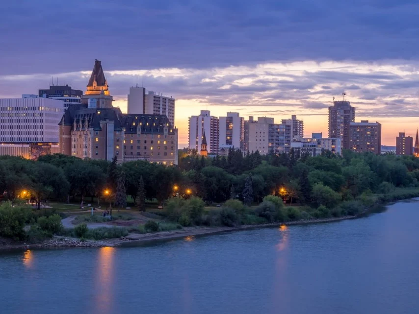 Uitzicht vanuit een rivieroeverpark op de skyline van Saskatoon bij zonsondergang met de torenspits van een kasteelachtig hotel tussen de gebouwen