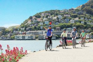Groep fietsers rijdt langs het water met op de achtergrond een heuvel vol kleurrijke huizen in Sausalito