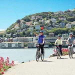 Een groep fietsers met helm rijdt langs het water in Sausalito, met een groen heuveldorp op de achtergrond