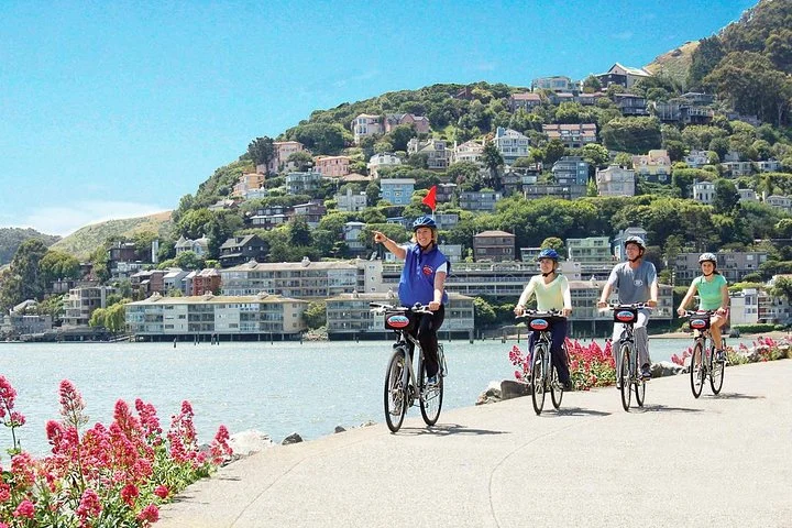 Een groep fietsers met helm rijdt langs het water in Sausalito, met een groen heuveldorp op de achtergrond