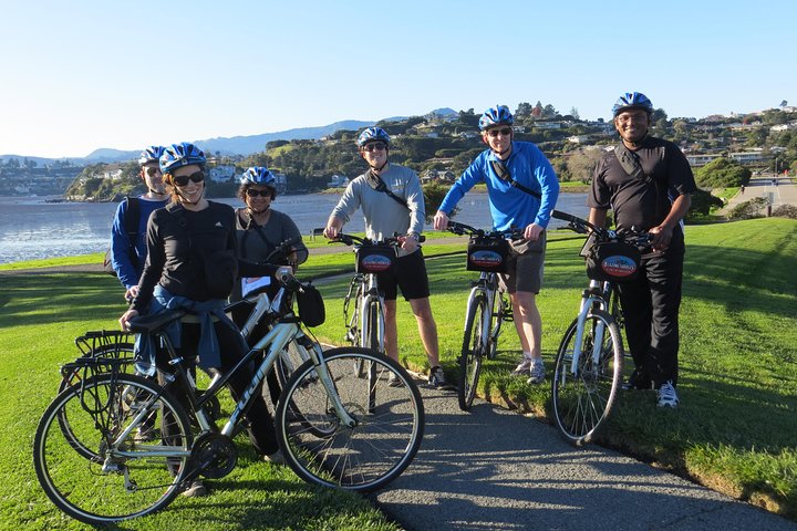 Zes fietsers met helmen staan met hun fietsen op het gras bij het water in Sausalito