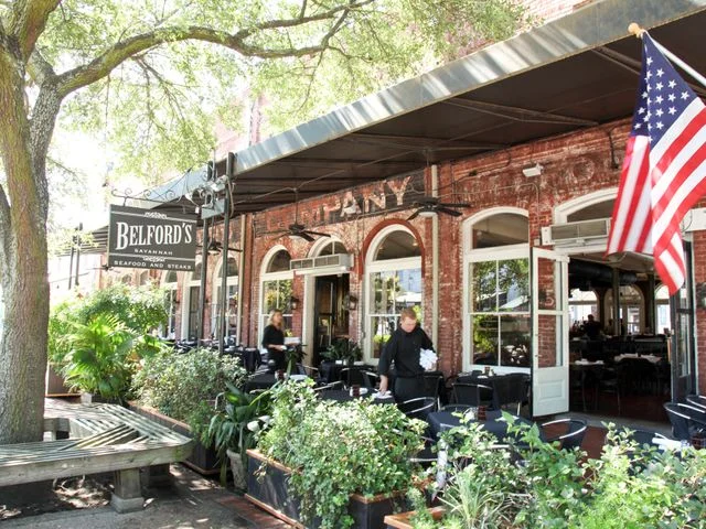 Buitenterras van restaurant Belford's in een bakstenen pand in Savannah, met tafels, planten en een Amerikaanse vlag aan de gevel