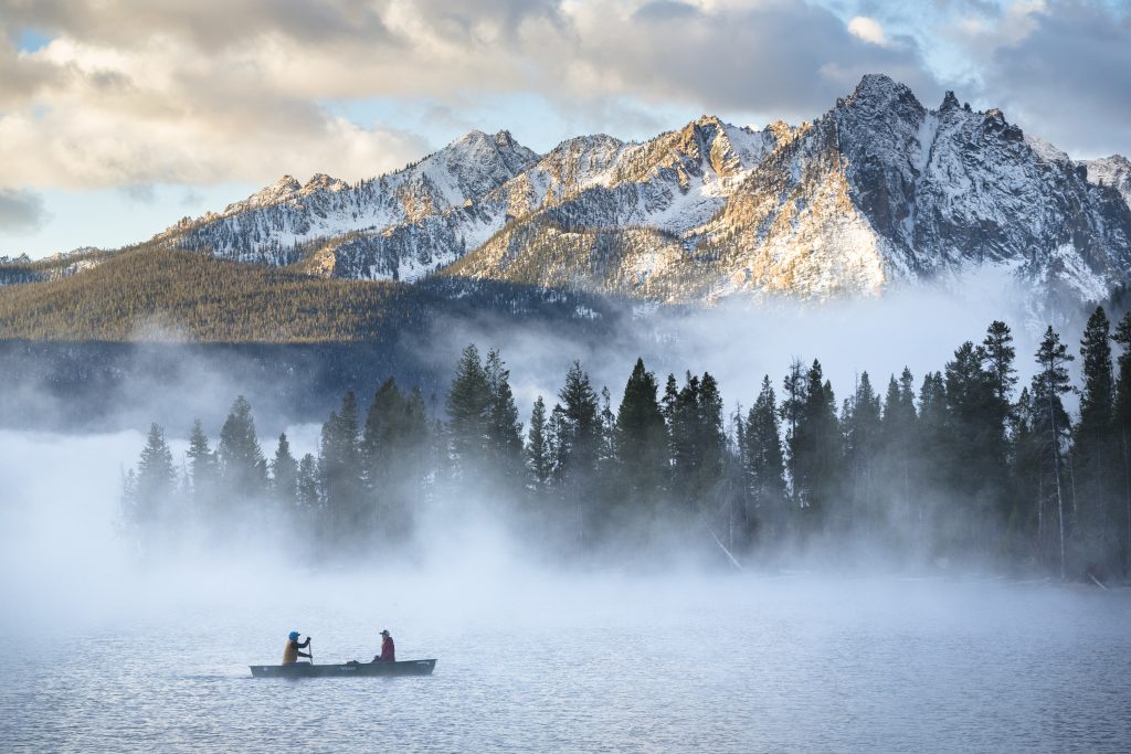 Twee mensen peddelen in een kano over een rustig meer met opstijgende mist, met dennenbos en besneeuwde bergtoppen erachter