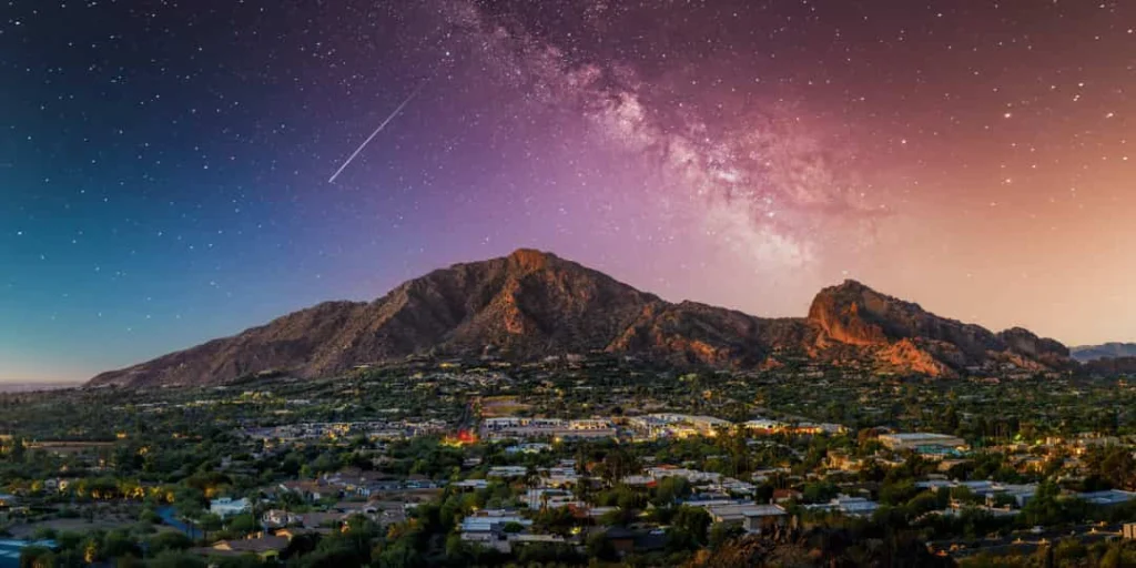 Silhouet van Camelback Mountain onder een sterrenhemel met de Melkweg en een vallende ster, met de verlichte stad eronder