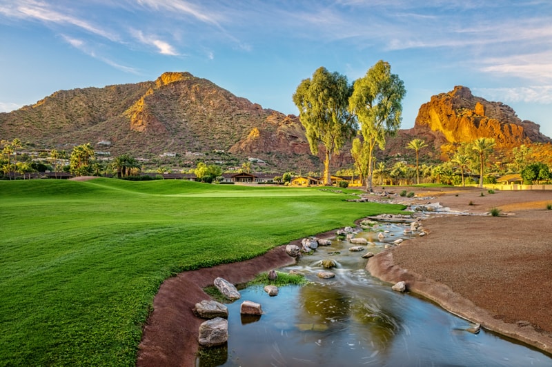Groene golfbaan met een kronkelend beekje over rotsen, bomen en op de achtergrond de kenmerkende rotsformatie van Camelback Mountain
