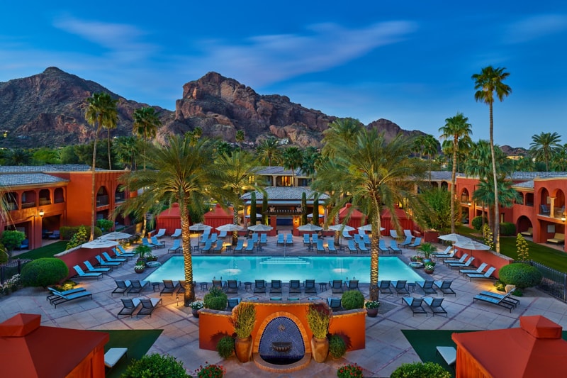 Groot zwembad met ligstoelen en parasols tussen palmbomen bij een luxe resort, met de rotsformatie van Camelback Mountain op de achtergrond in de avondschemering