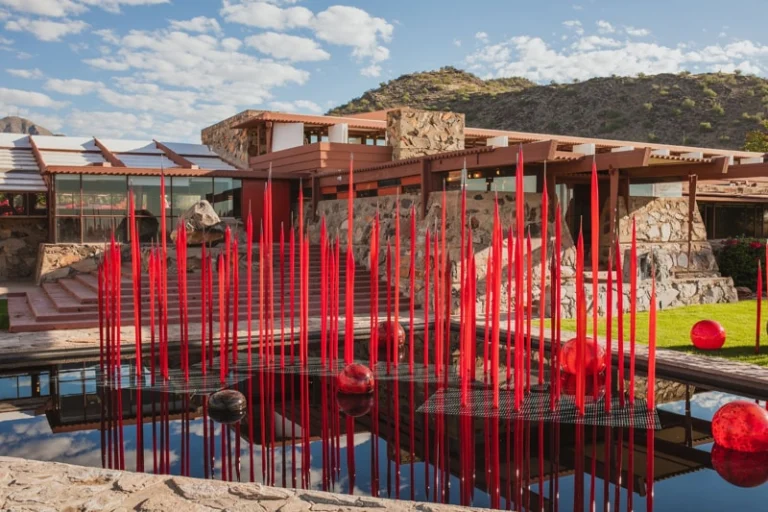 Installatie van hoge, rode glazen rietstengels op een grasveld voor het door Frank Lloyd Wright ontworpen gebouw Taliesin West, met bergen op de achtergrond