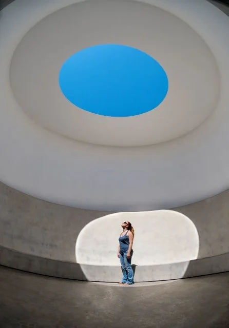 Bezoeker staat in een ronde, witte ruimte met een groot rond gat in het plafond waardoor de blauwe lucht zichtbaar is