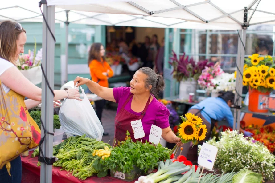 Marktvrouw overhandigt een tas met verse groenten aan een klant bij een kraam vol zonnebloemen en groenten op Pike Place Market in Seattle