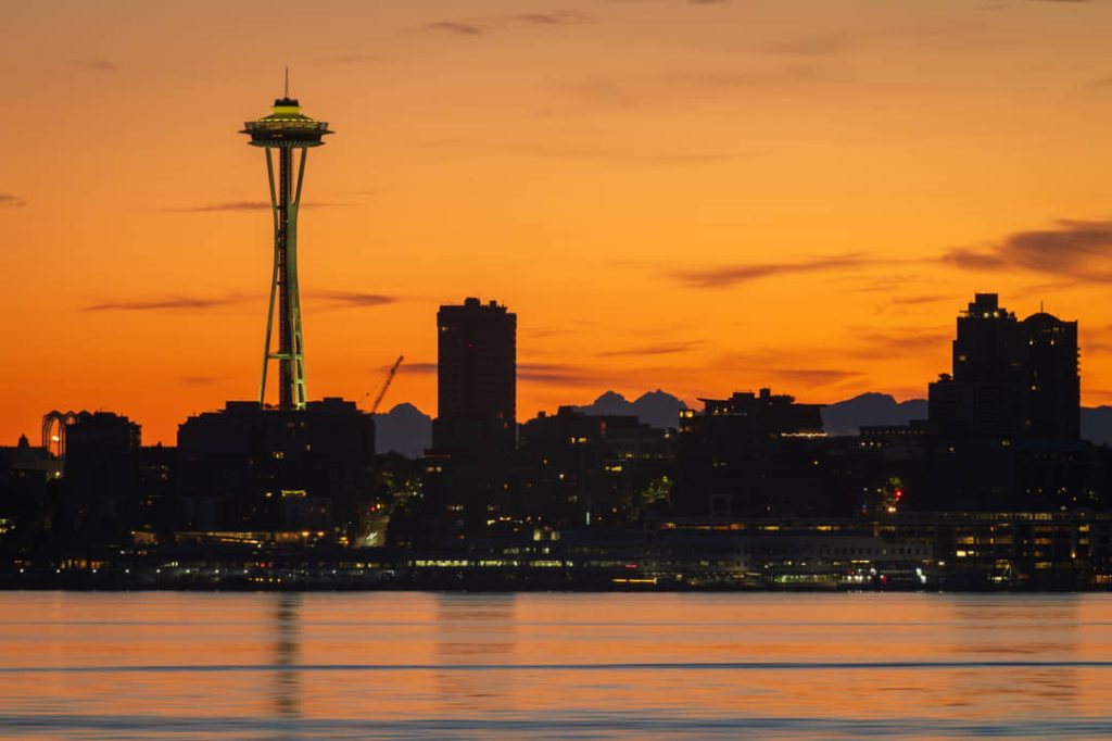 Silhouet van de Space Needle en de wolkenkrabbers van Seattle tegen een oranje avondlucht met het water op de voorgrond