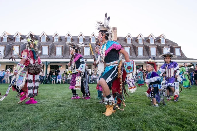 Groep dansers in kleurrijke traditionele kleding met veren en kralen dansen op het gazon voor een groot houten hotel met dakkapellen